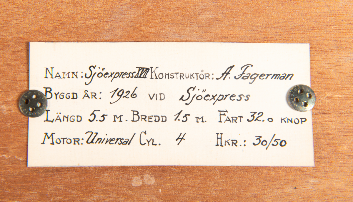 Halvmodell av motorbåt, racer. Byggd i block av trä visande styrbords sida. Skrov av mahogny med kopparfärgad botten. Monterad på svart polerad platta av mahogny med ljus ram.