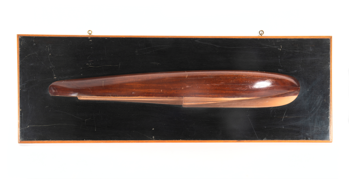 Halvmodell av motorbåt, racer. Byggd i block av trä visande styrbords sida. Skrov av mahogny med kopparfärgad botten. Monterad på svart polerad platta av mahogny med ljus ram.