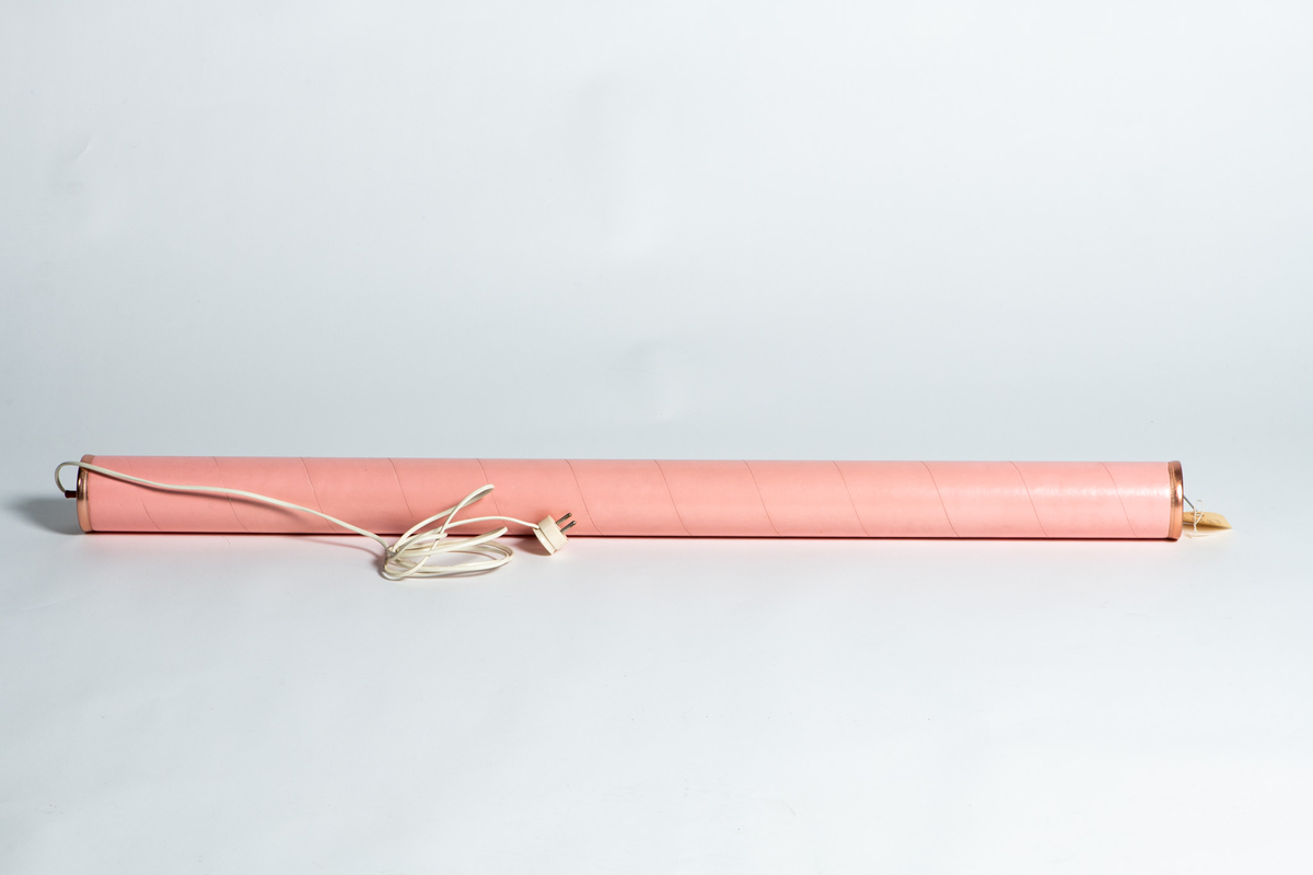 Elektrisk sengevarmer "Don Juan". Sylinderform. Overflaten belagt med rosa plastlignende stoff, silkematt. Hempe for opphengning på vegg, den andre med feste for ledning  av hvit  plast  med støpsel.  
