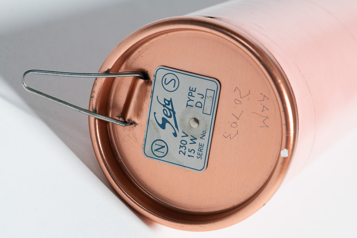 Elektrisk sengevarmer "Don Juan". Sylinderform. Overflaten belagt med rosa plastlignende stoff, silkematt. Hempe for opphengning på vegg, den andre med feste for ledning  av hvit  plast  med støpsel.  
