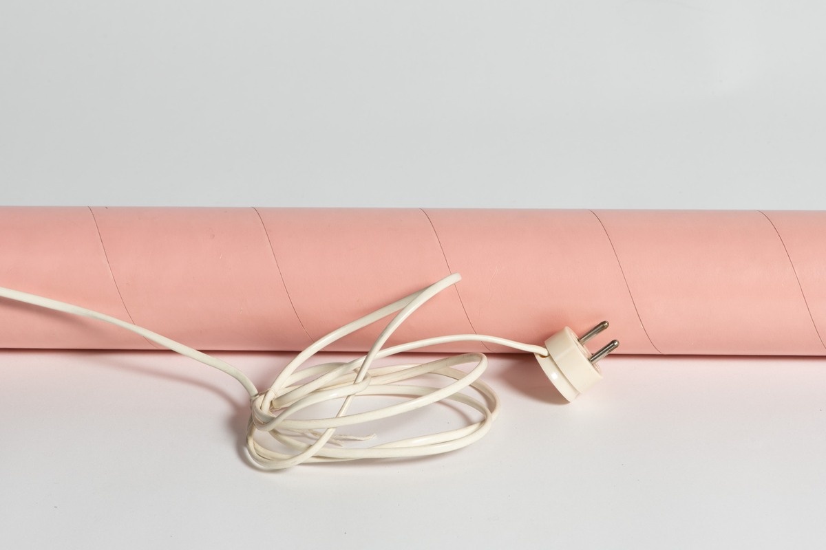 Elektrisk sengevarmer "Don Juan". Sylinderform. Overflaten belagt med rosa plastlignende stoff, silkematt. Hempe for opphengning på vegg, den andre med feste for ledning  av hvit  plast  med støpsel.  
