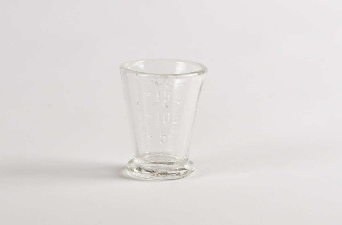 Lite målebeger i glass. Flat stett som skråner litt ut nederst. Glasset skråner jevnt oppover fra bunnen. Sirkelrundt åpning på toppen. I glasset er det støpt inn målegraderinger: - TS, - BS, og - Sps.
