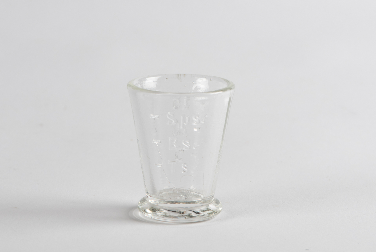 Lite målebeger i glass. Flat stett som skråner litt ut nederst. Glasset skråner jevnt oppover fra bunnen. Sirkelrundt åpning på toppen. I glasset er det støpt inn målegraderinger: - TS, - BS, og - Sps.