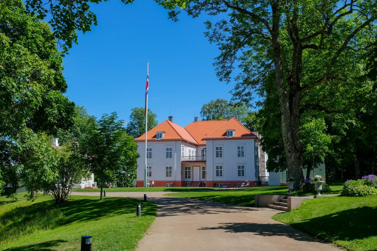 Eidsvollsbygningen sett forfra en sommerdag, med flaggstang, blå himmel og grønne trær rundt.