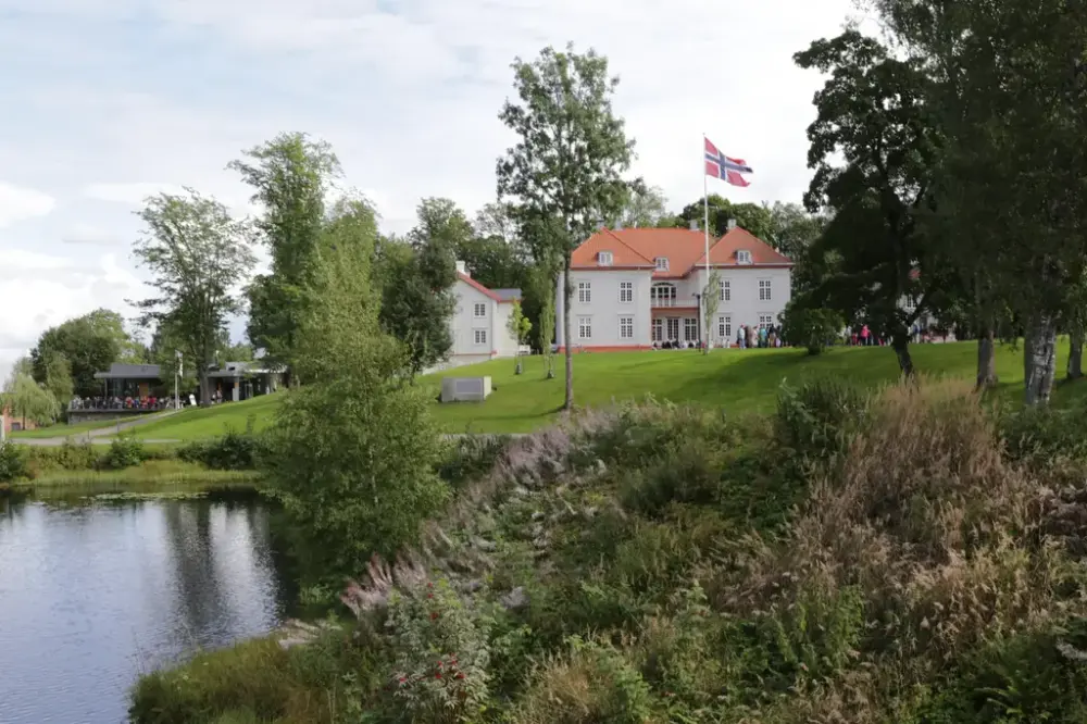 Eidsvollsbygningen sett fra hagesiden, med norsk flagg vaiende foran bygningen, vannet til Andelva og grønt landskap rundt.
