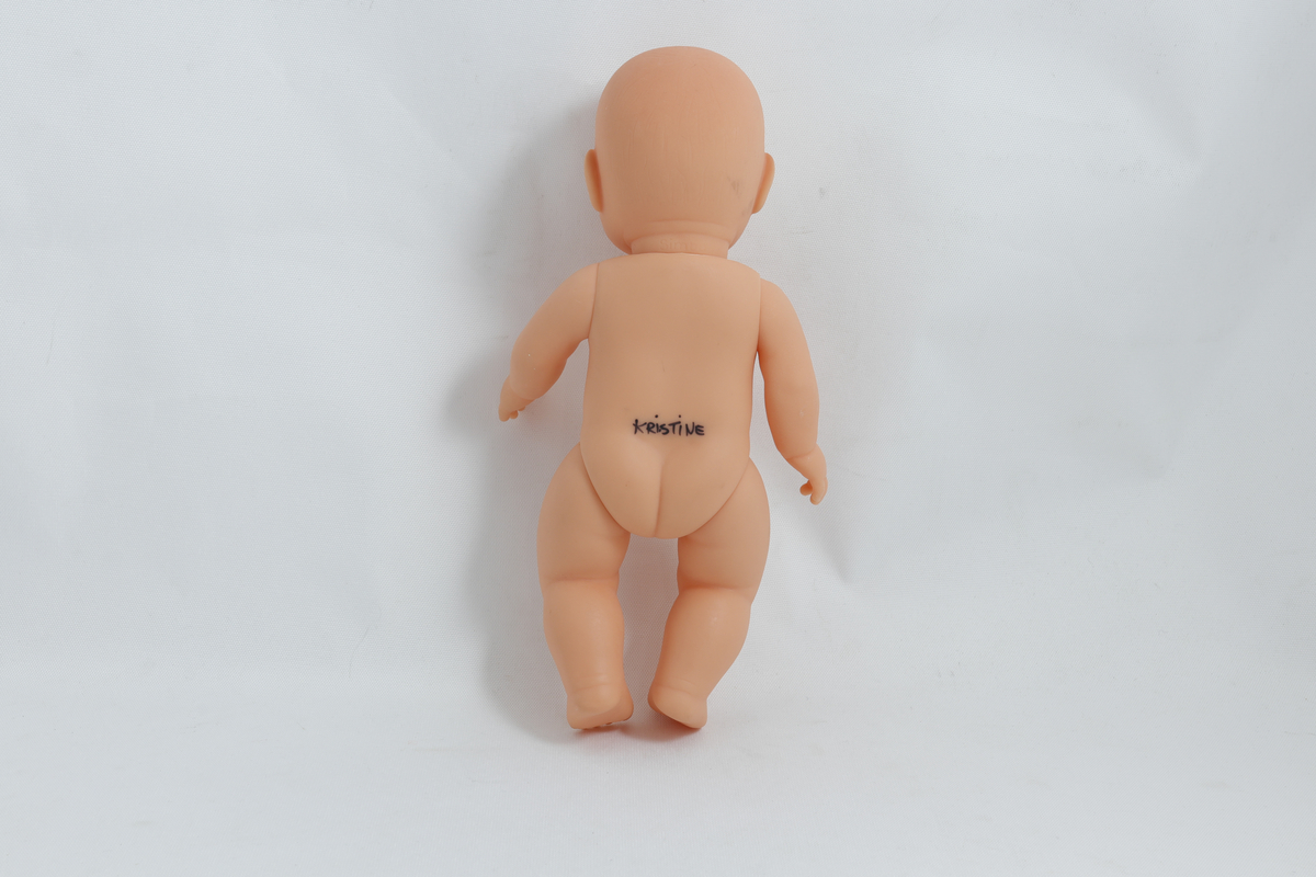 Babydukke, uten klær. Det er skrevet "Kristine" med tusj nederst på ryggen på dukka. 