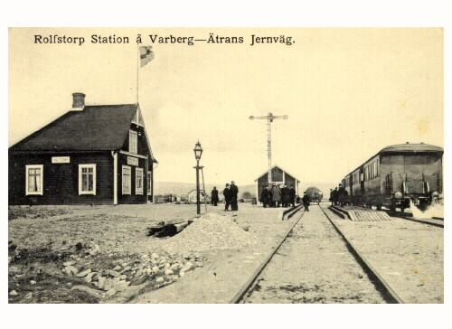 Vykort, "Rolfstorp Station å Varberg-Ätrans Jernväg. Järnvägen invigdes 1911 och på bilden ses banområdet där en klunga personer står vid semaforen. Ett tåg står inne vid perrongen. Stationshusets gavel pryds av en hissad flagga.