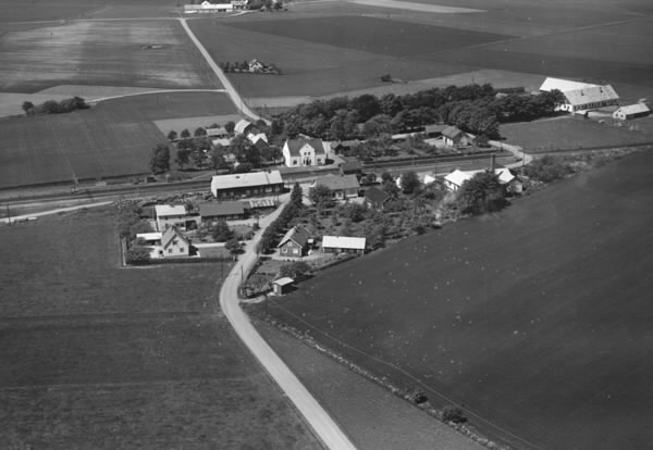 "Flygfoto över Brännarp" i Rävinge. Här startade 1938 Hallands Fruktindustri (HAFI) som fortfarande är verksamt. Där järnvägen korsar landsvägen ligger stationsbyggnaden, vilken fick detta utseende 1921 efter en kraftig ombyggnad. Stationen omges av ett fåtal villor och några lantgårdar med  ekonomibyggnader och omgivande odlingslandskap.
