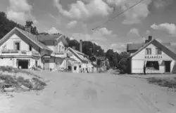 Stationssamhället i Vessigebro. Gata kantad av affärsbyggnader, bland annat livsmedelsaffär och fiskaffär. Cyklar står parkerade längs gatan. Skyltar på byggnaderna: "Livsmedel", "BLOMMOR", "GRANGÉUS" och "FISKAFFÄR". Vägskylt med texten: "Vessigebro stn".