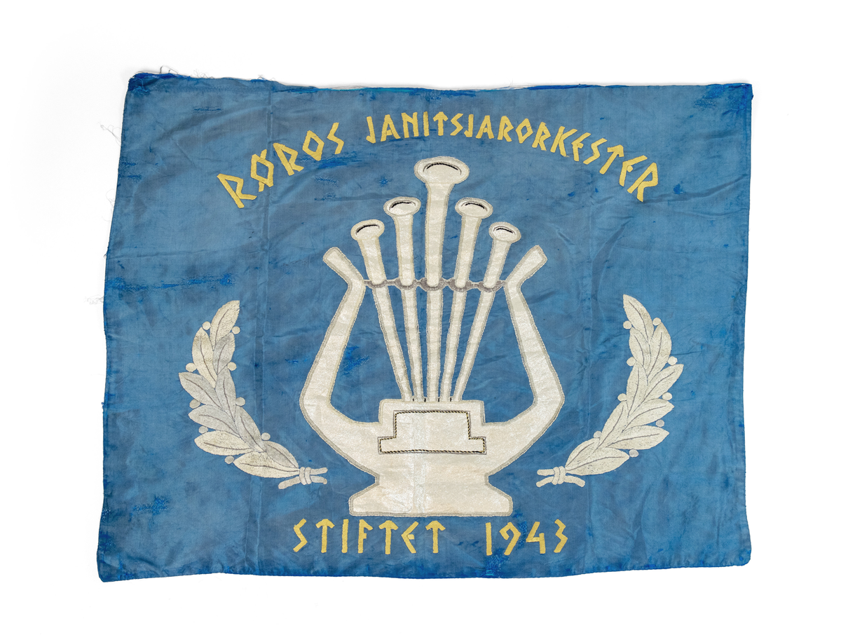 Fane i silke etter Røros Janitsjarorkester. Påbrodert motiv som illustrerer bladkrans. Applikert motiv som illustrerer lyre. Påskrift: Røros Janitsjarorkester stiftet 1943 brodert med gult bomullsgarn. Reparert og stoppet flere steder. Tilhørende posement; en frynsekant og tre sett med snorer. 
