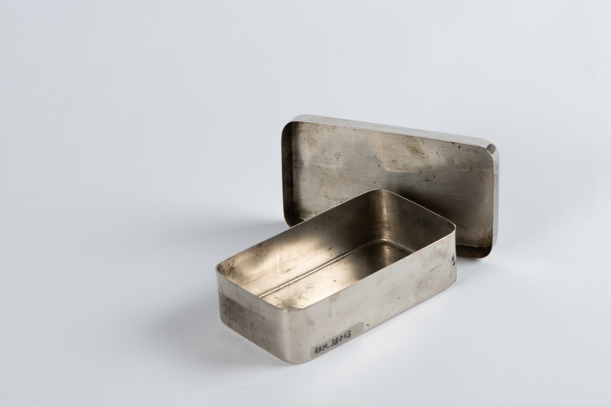 Rektangulær metalleske med lokk. Avrundede hjørner. En teipbit på oversiden som virker å være fra når den ble brukt. To hvite klistremerker klistret på hver sin side i etterkant. Esken er tom.