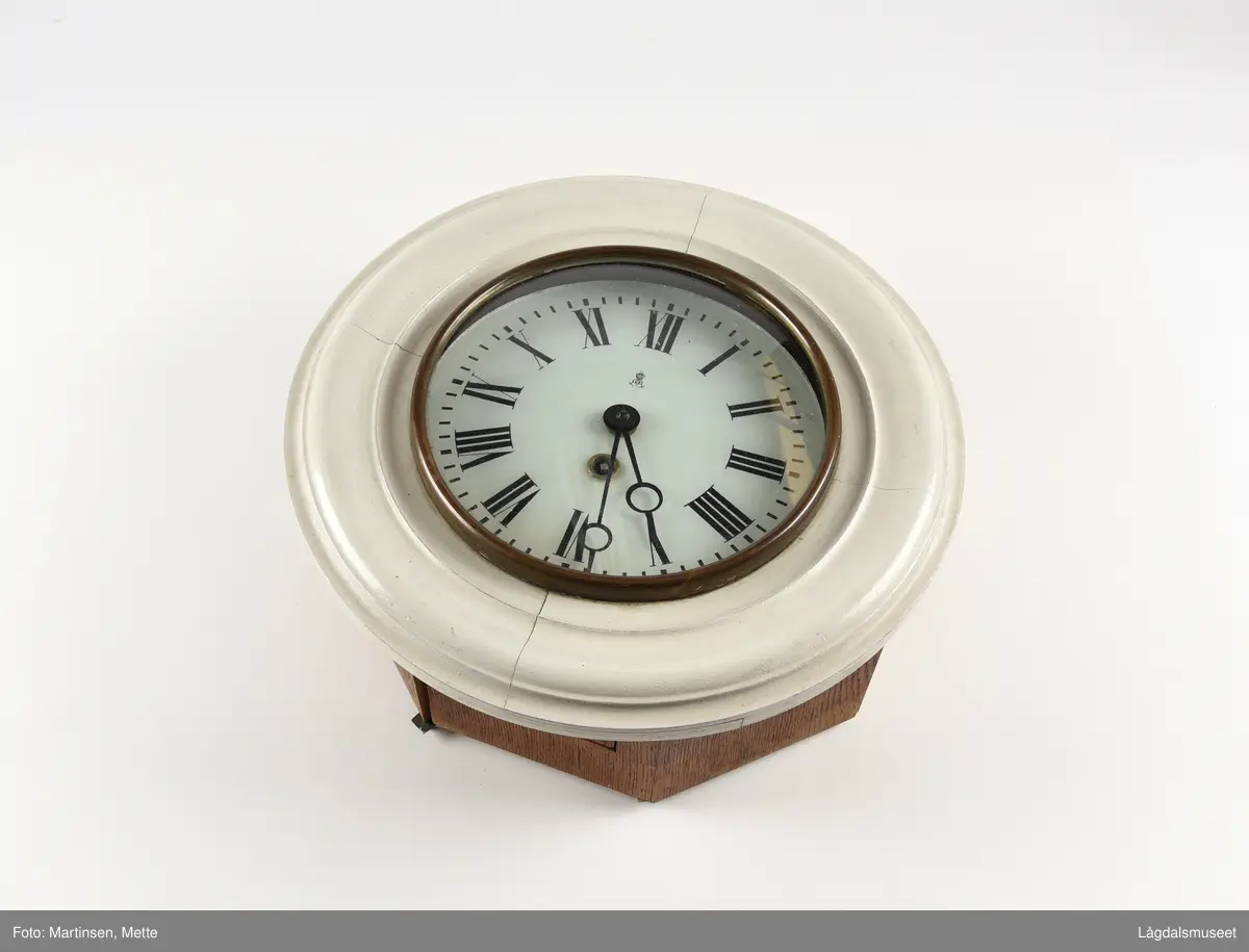 Sirkelformet urskive og deksel, 8-kantet kasse. Profilerte lister rundt deksel. Liten luke som kan åpnes i bunnsiden av kassen. På urskiven bokstavene GB (Gustav Becker) samt anker og krone. 