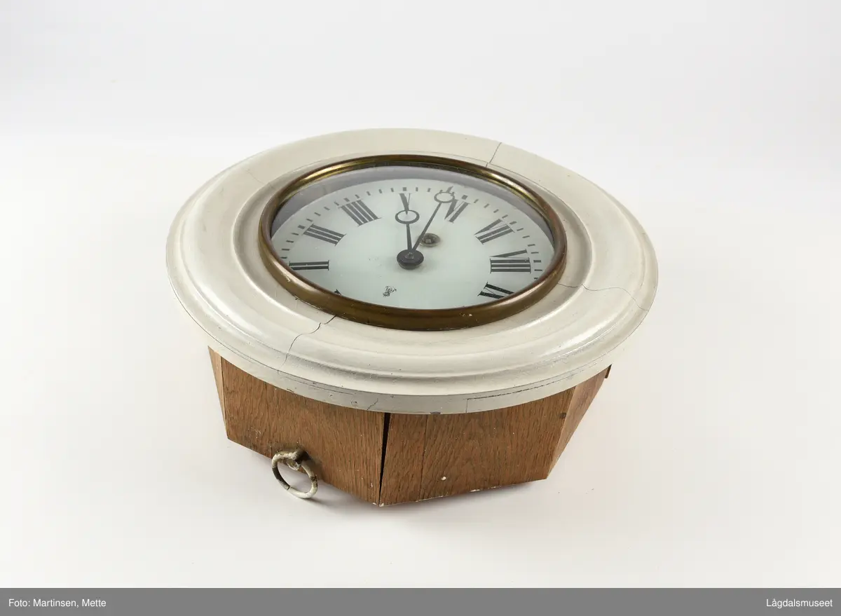 Sirkelformet urskive og deksel, 8-kantet kasse. Profilerte lister rundt deksel. Liten luke som kan åpnes i bunnsiden av kassen. På urskiven bokstavene GB (Gustav Becker) samt anker og krone. 