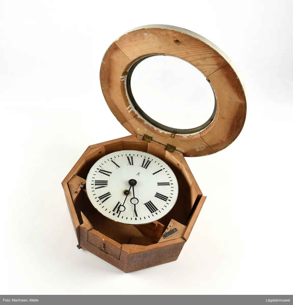 Sirkelformet urskive og deksel, 8-kantet kasse. Profilerte lister rundt deksel. Liten luke som kan åpnes i bunnsiden av kassen. På urskiven bokstavene GB (Gustav Becker) samt anker og krone. 