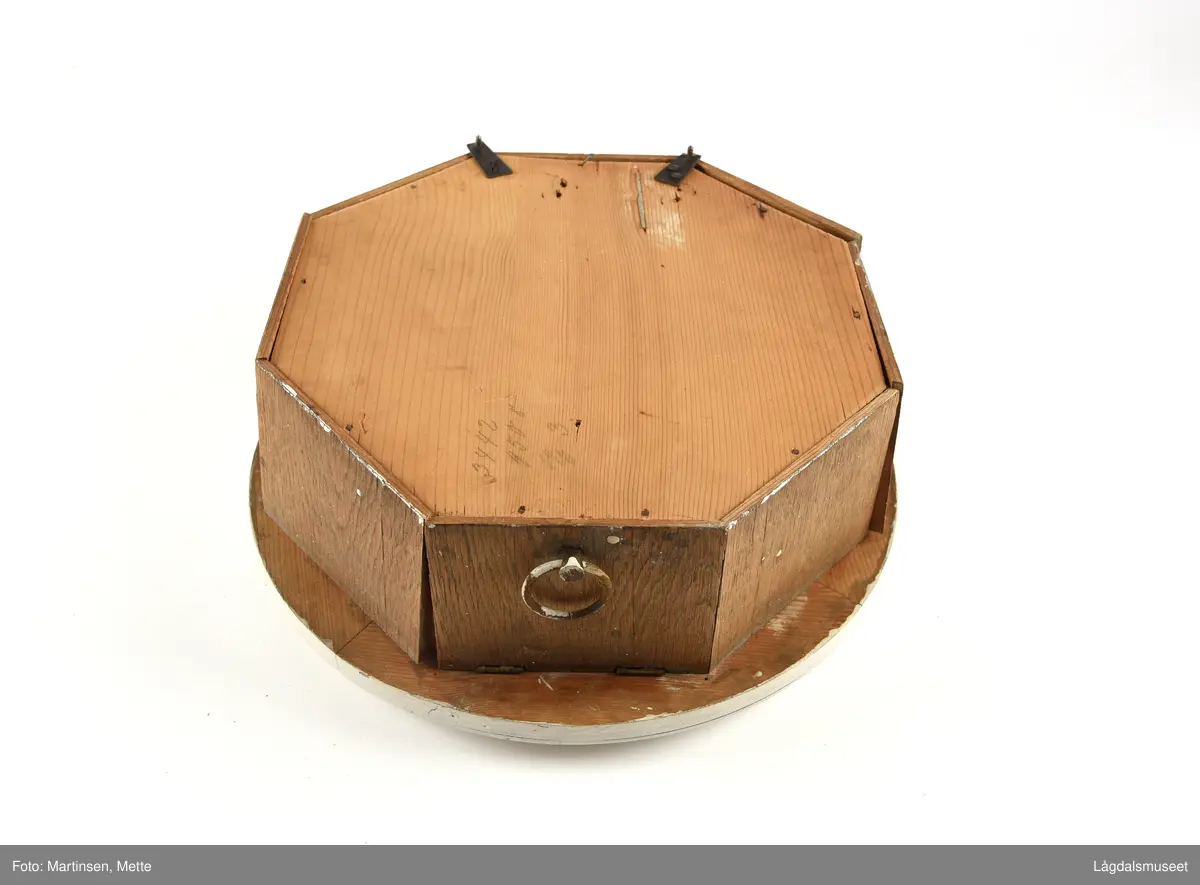 Sirkelformet urskive og deksel, 8-kantet kasse. Profilerte lister rundt deksel. Liten luke som kan åpnes i bunnsiden av kassen. På urskiven bokstavene GB (Gustav Becker) samt anker og krone. 