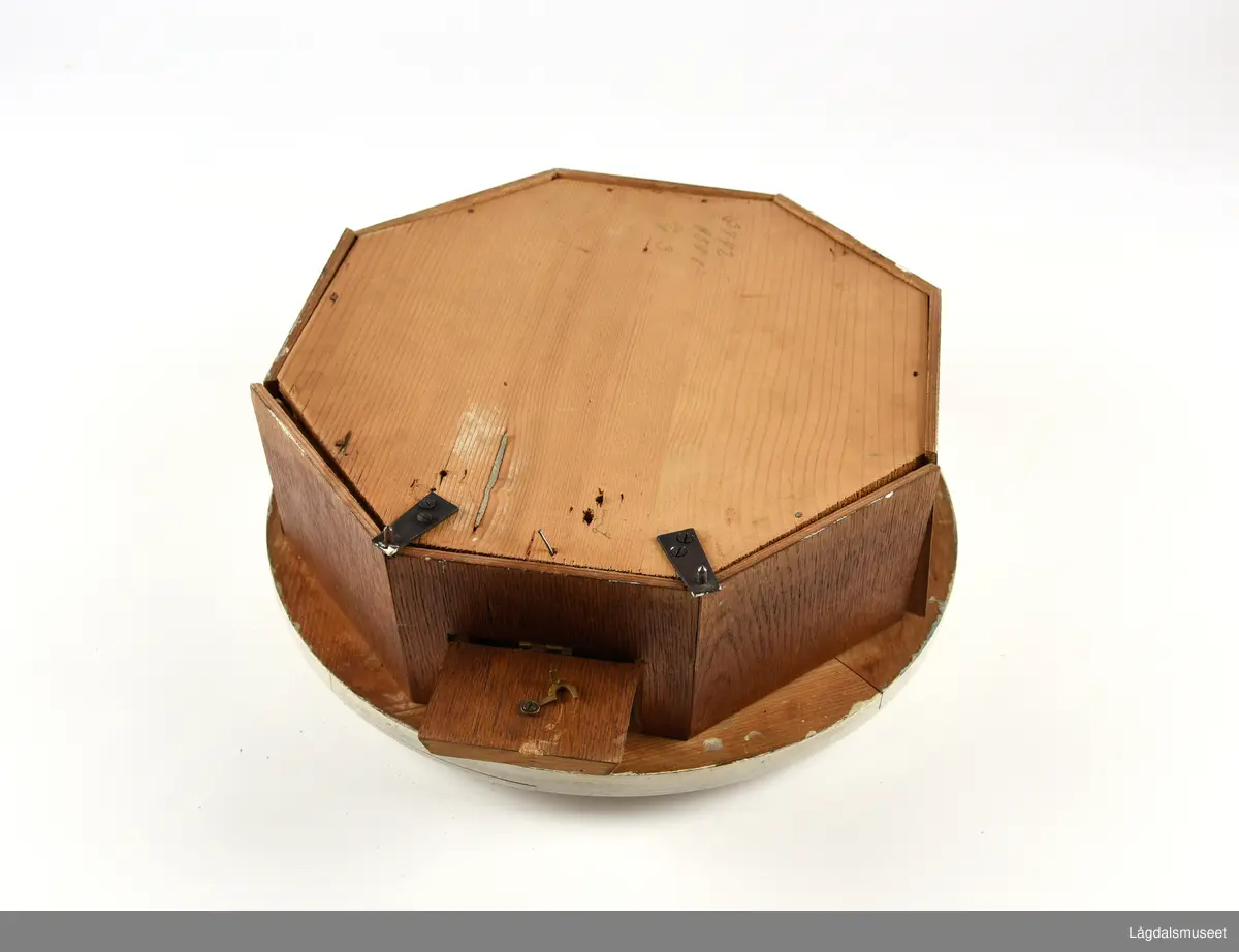 Sirkelformet urskive og deksel, 8-kantet kasse. Profilerte lister rundt deksel. Liten luke som kan åpnes i bunnsiden av kassen. På urskiven bokstavene GB (Gustav Becker) samt anker og krone. 