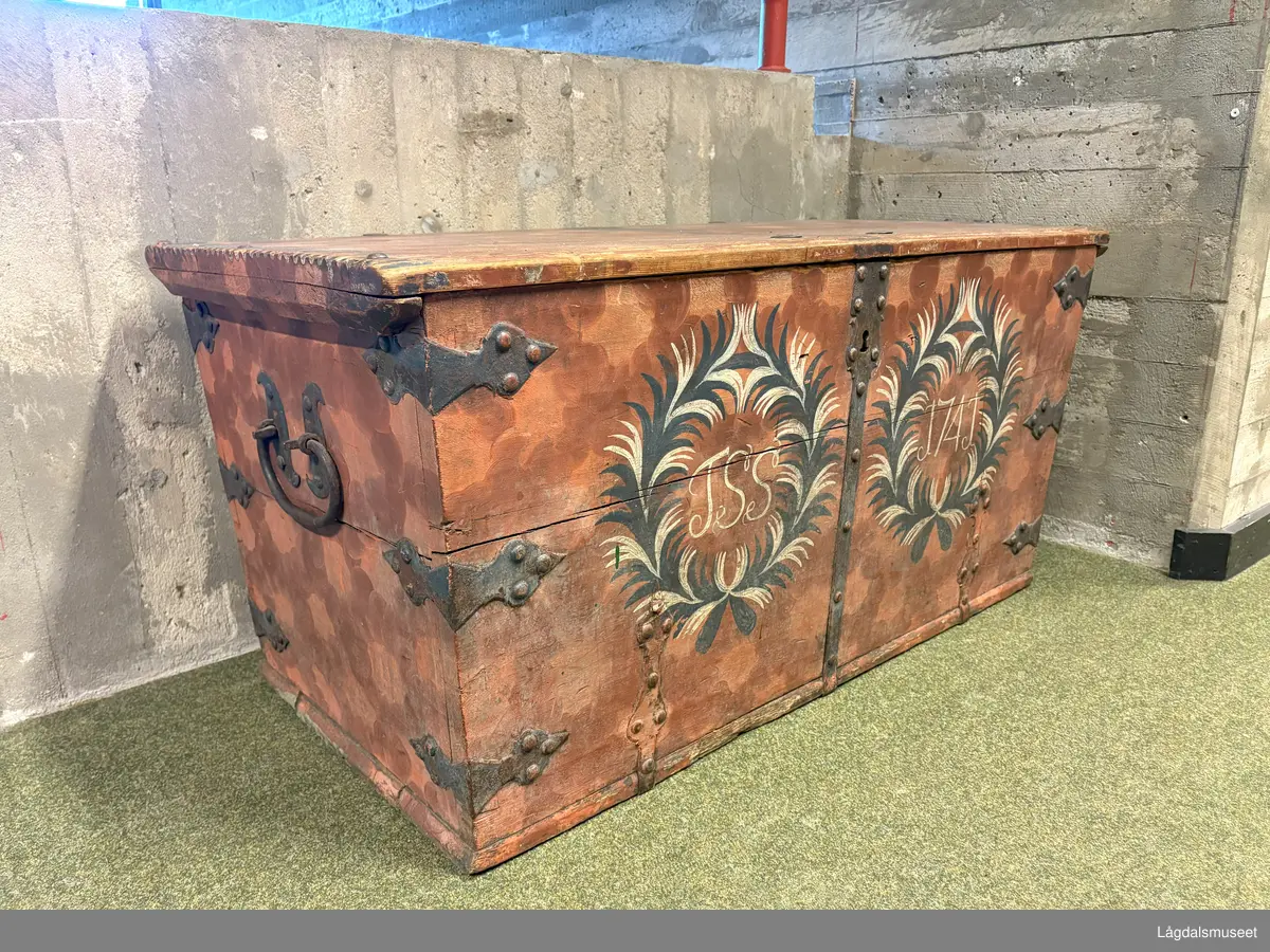 Dekorert kiste i tre med flatt lokk og leddik til høyre. Tre hjørnebeslag i jern på hvert hjørne, samt et foran på kista. Påmalt etterligning av beslag på lokket. 
