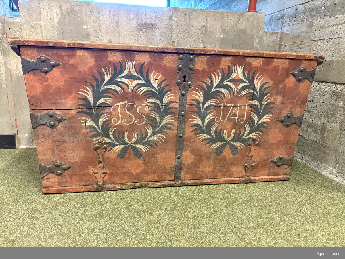 Dekorert kiste i tre med flatt lokk og leddik til høyre. Tre hjørnebeslag i jern på hvert hjørne, samt et foran på kista. Påmalt etterligning av beslag på lokket. 