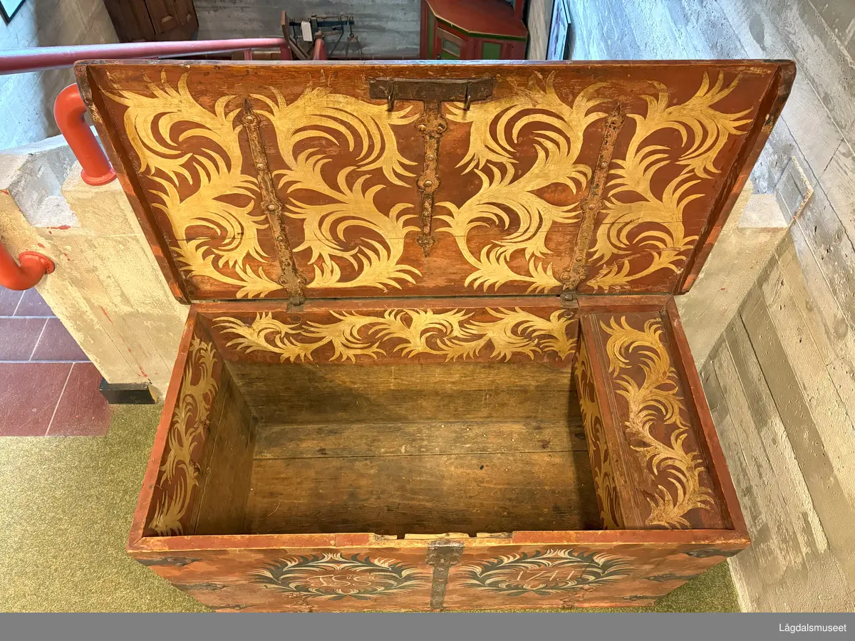 Dekorert kiste i tre med flatt lokk og leddik til høyre. Tre hjørnebeslag i jern på hvert hjørne, samt et foran på kista. Påmalt etterligning av beslag på lokket. 