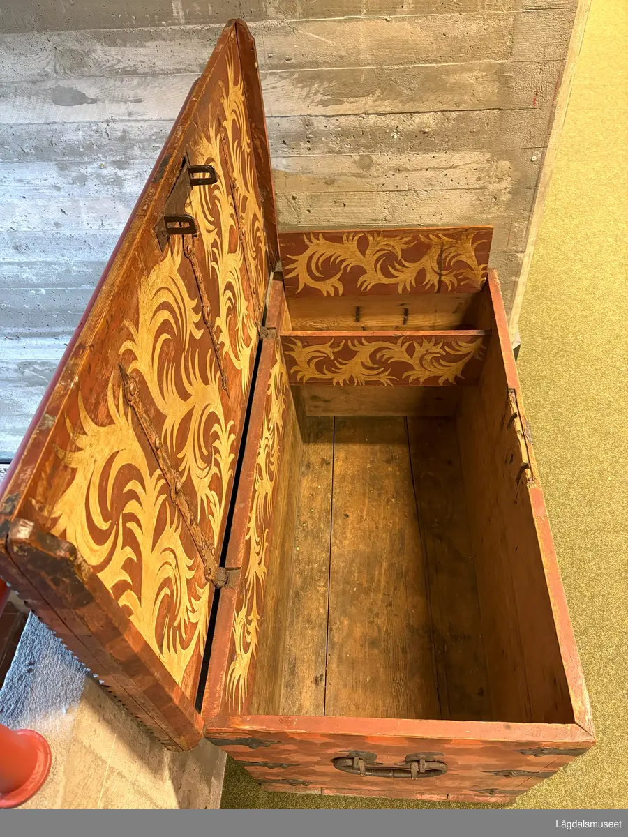 Dekorert kiste i tre med flatt lokk og leddik til høyre. Tre hjørnebeslag i jern på hvert hjørne, samt et foran på kista. Påmalt etterligning av beslag på lokket. 