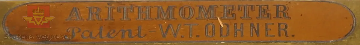 Mekanisk regnemaskin laget av metall som har et sveivehåndtak. Regnemaskinen sitter i en tre kasse med messinglås og trehåndtak. Regnemaskinen er gullfarget og svart, og er dekorert med logo. Sveivehåndtaker er sølv og brun farget og laget av tre. Inne i kisten henger en lapp. Det ligger også en liten nøkkel i kisten som hører til låsen. 