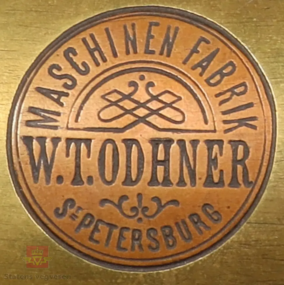 Mekanisk regnemaskin laget av metall som har et sveivehåndtak. Regnemaskinen sitter i en tre kasse med messinglås og trehåndtak. Regnemaskinen er gullfarget og svart, og er dekorert med logo. Sveivehåndtaker er sølv og brun farget og laget av tre. Inne i kisten henger en lapp. Det ligger også en liten nøkkel i kisten som hører til låsen. 