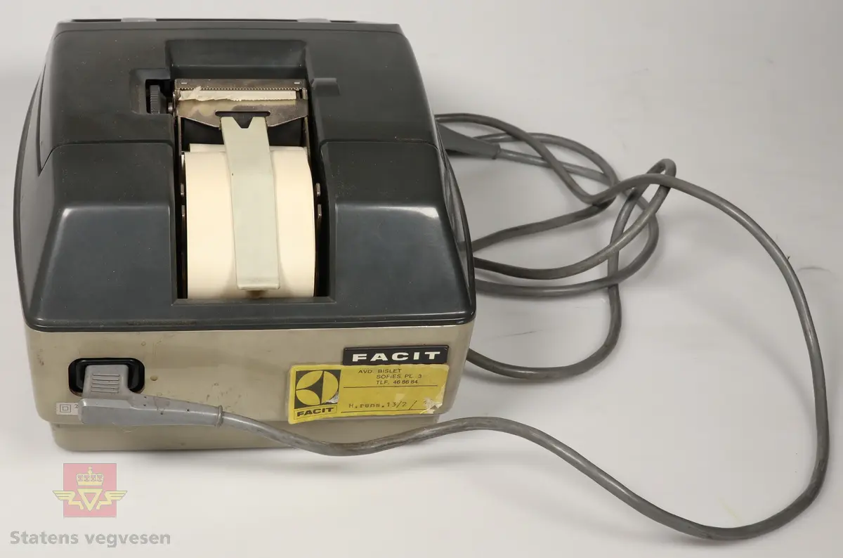 Elektrisk regnemaskin i varierte gråtoner. Stor, rektangulær form. Den har en papirrull på toppen. Maskinens taster er mørkegrå, lyse grå, hvite og røde. Den har en lysegrå ledning. 