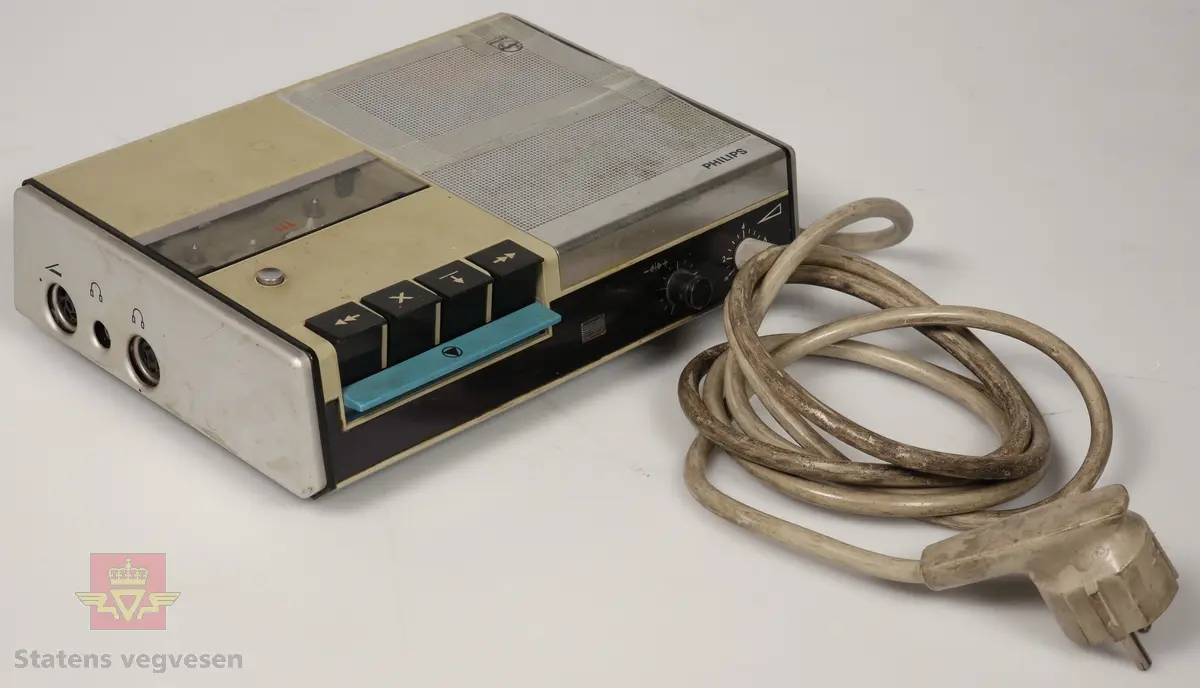 Dikteringsmaskin som er lyse grå, rektangulær og har fire svarte knapper på høyre siden, med en blå spak. Har svart panel for og et sølv-grått panel på toppen. Har PHILIPS markert på toppen. En gjennomsiktig luke på toppen med en sølv knapp som gjør at luken åpnes når den trykkes på. Den har en hvit ledning.
Gjenstander ligner på NVM 00-G-00082, men har flere forskjeller. 