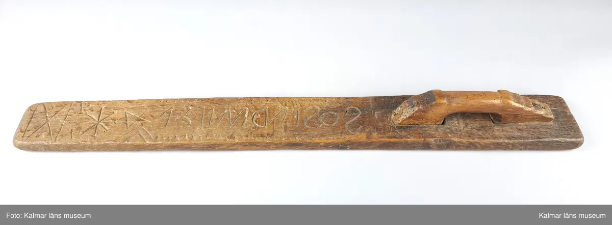 KLM 6438. Mangelbräde. Tillverkat av ek. Platt. Längs större delen av kanten vinkelband. Ytan har grovt inskurna tecken. Inskription bakvänd: SOS LAD AN 1734. Smalt men enkelt snidat handtag utan hästhuvud.