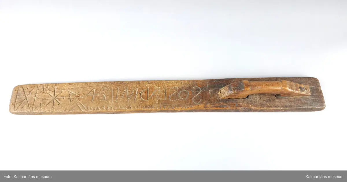 KLM 6438. Mangelbräde. Tillverkat av ek. Platt. Längs större delen av kanten vinkelband. Ytan har grovt inskurna tecken. Inskription bakvänd: SOS LAD AN 1734. Smalt men enkelt snidat handtag utan hästhuvud.