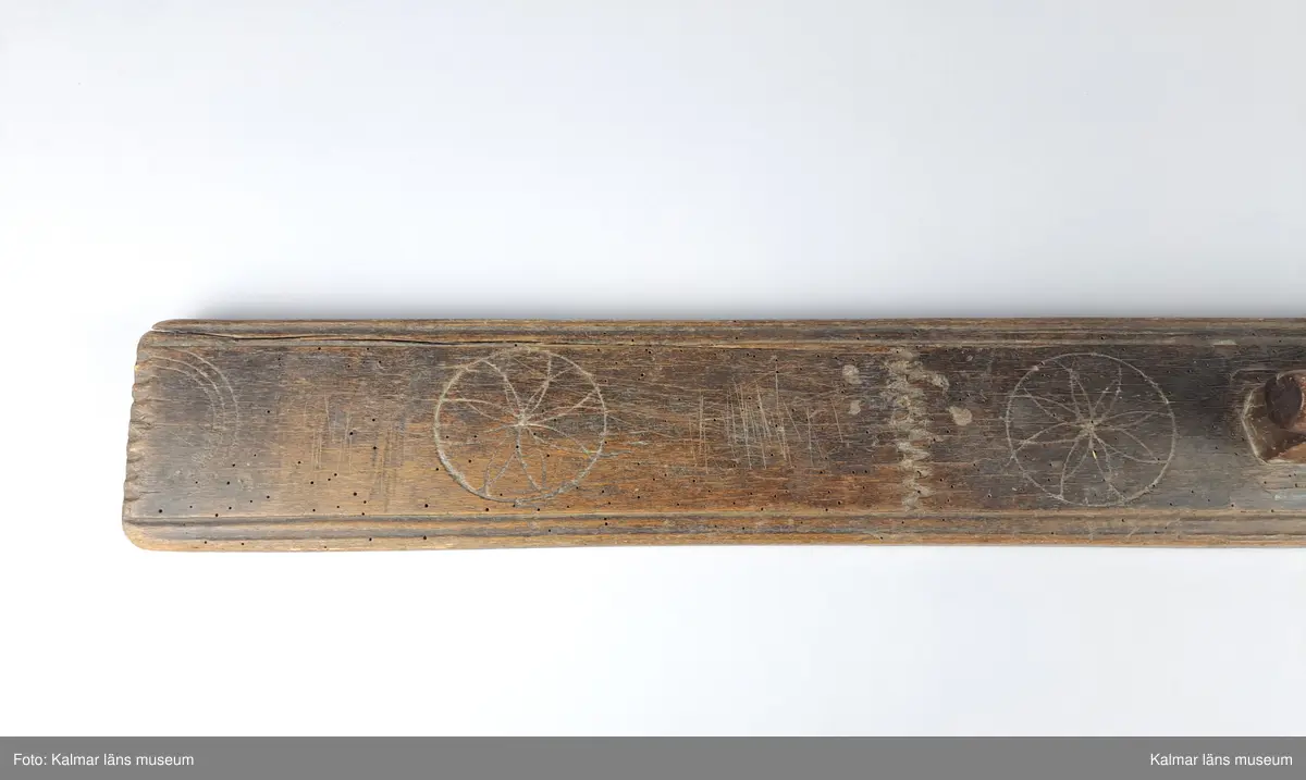 KLM 10521:1. Mangelbräde. Tillverkat av trä. Enkelt utsnidat med kälade kanter. Dekor i form av cirklar med blommönster. Enkelt handtag.