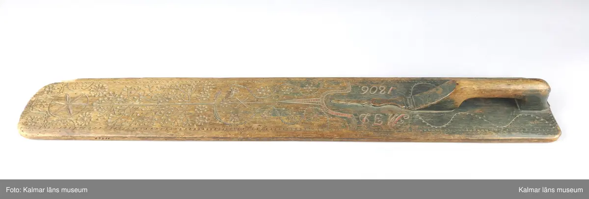 KLM 11552. Mangelbräde. Av trä. Med snidat handtag. Rikt snidat, uddmönster i kanterna, slingor, ett träd, tornliknade utsmyckning, en större blomma och blommor som består av streck och punkter. Inskrift: M.J.D. 1806.