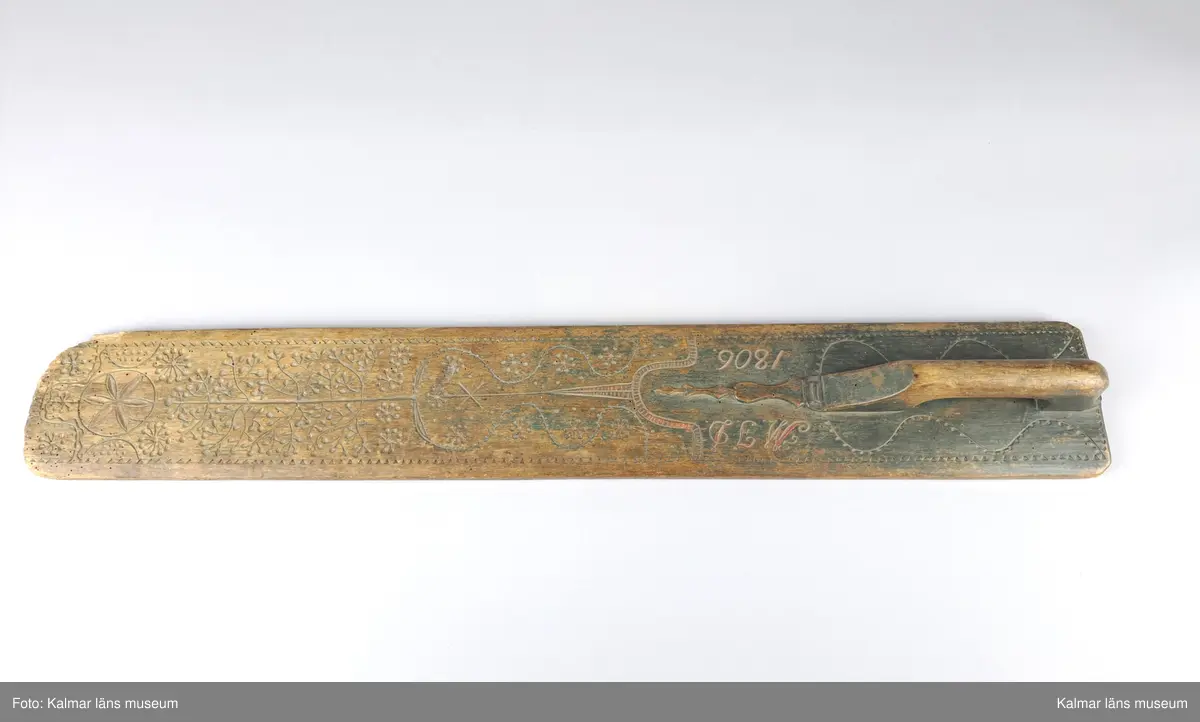 KLM 11552. Mangelbräde. Av trä. Med snidat handtag. Rikt snidat, uddmönster i kanterna, slingor, ett träd, tornliknade utsmyckning, en större blomma och blommor som består av streck och punkter. Inskrift: M.J.D. 1806.