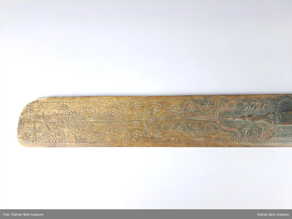 KLM 11552. Mangelbräde. Av trä. Med snidat handtag. Rikt snidat, uddmönster i kanterna, slingor, ett träd, tornliknade utsmyckning, en större blomma och blommor som består av streck och punkter. Inskrift: M.J.D. 1806.