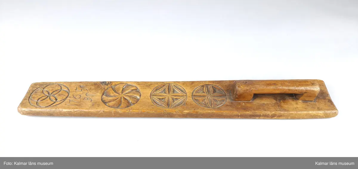 KLM 13798:1. Mangelbräde. Tillverkat av ek med snidad dekor i karvsnittsrosetter. Snidat handtag. Brännstämpel på brädets undersida: J.G.HEDIN. Inskrift ovansida: 1765 PT.