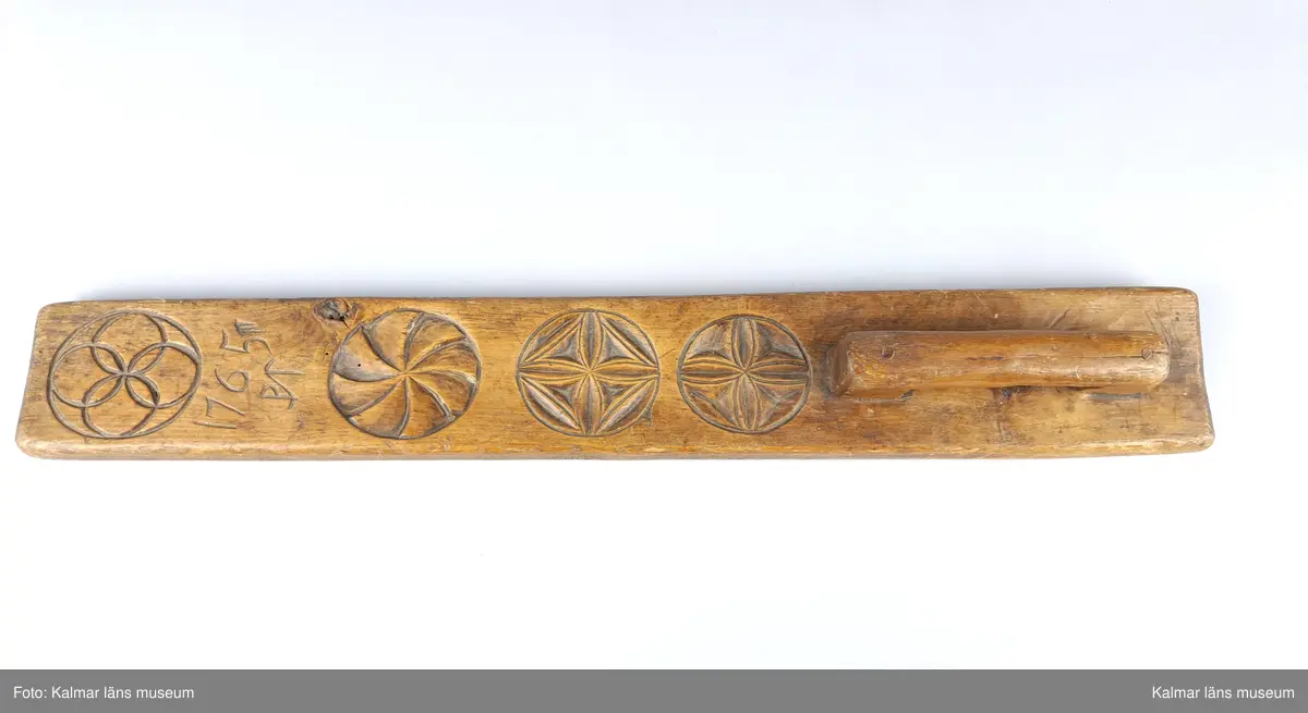 KLM 13798:1. Mangelbräde. Tillverkat av ek med snidad dekor i karvsnittsrosetter. Snidat handtag. Brännstämpel på brädets undersida: J.G.HEDIN. Inskrift ovansida: 1765 PT.