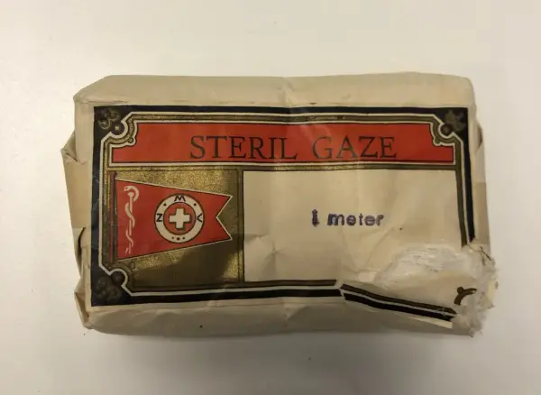 Bildet viser en pakke i gråpapir og med et trykk i rødt og sort og gull på den ene siden. Det står skrevet STERIL GAZE og "i meter"