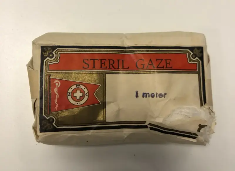 Bildet viser en pakke i gråpapir og med et trykk i rødt og sort og gull på den ene siden. Det står skrevet STERIL GAZE og "i meter"