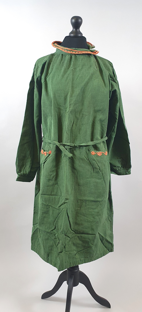 Grønn kjole med lange ermer. Krage med trykknapper på venstre side og med oransje broderier. To innsydde lommer foran, med samme broderier som på kragen. Belte i livet.