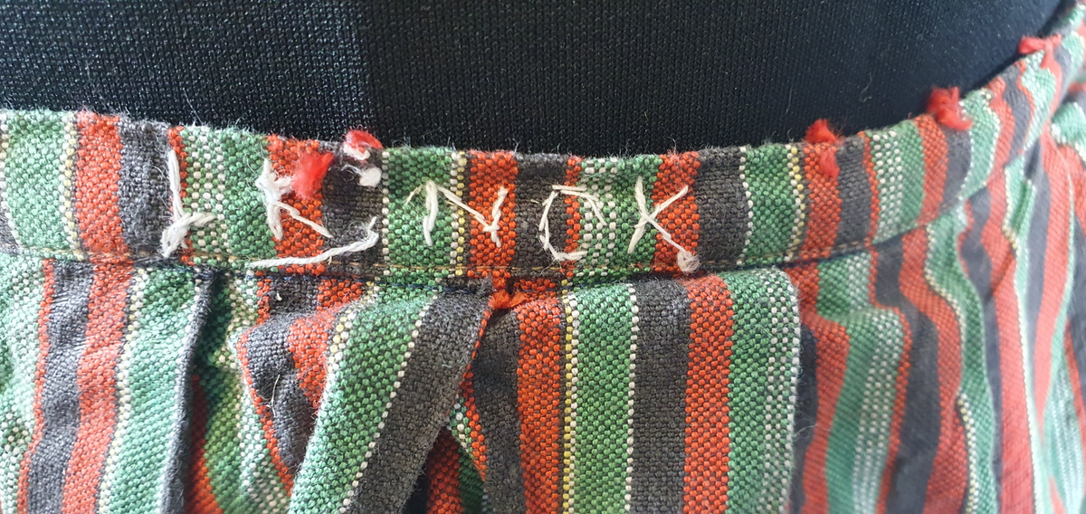 Skjørt med smale, vertikale striper i rødt, grønt, svart og hvitt. Smal linning i livet. God vidde. Brodert navn i linningen.
