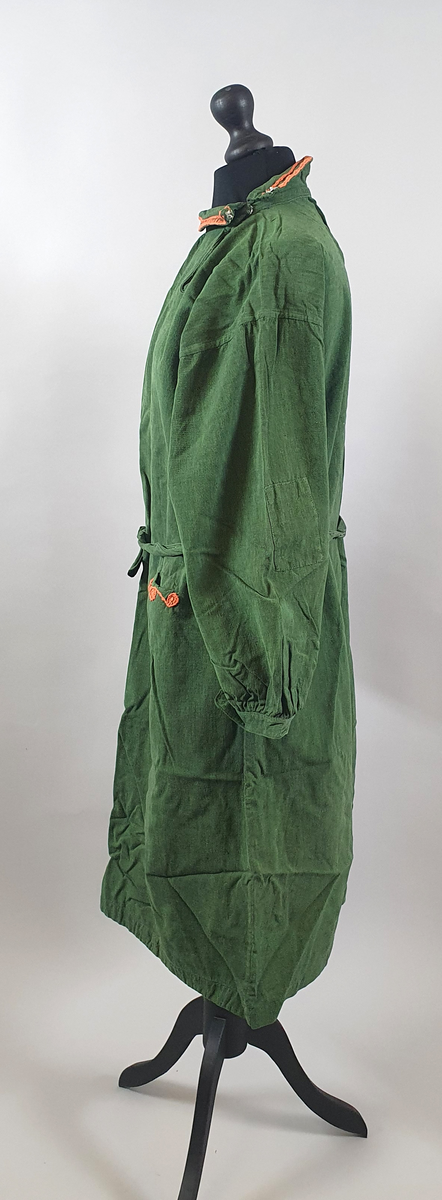 Grønn kjole med lange ermer. Krage med trykknapper på venstre side og med oransje broderier. To innsydde lommer foran, med samme broderier som på kragen. Belte i livet.