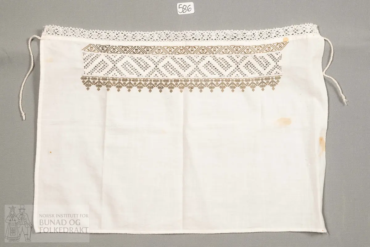 Hylk, firkanta stoffstykke. Sydd av kvit bomullslerret. Falda for hand. Broderi sydd med uttrekkssaum. På kvar side av denne er brodert med svartbrun silketråd, korssting og holbeinsting. Påsydd knyteband i framkant.