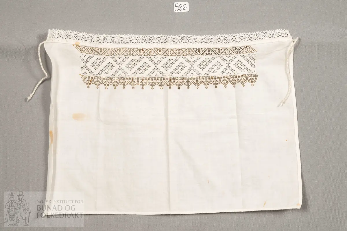 Hylk, firkanta stoffstykke. Sydd av kvit bomullslerret. Falda for hand. Broderi sydd med uttrekkssaum. På kvar side av denne er brodert med svartbrun silketråd, korssting og holbeinsting. Påsydd knyteband i framkant.