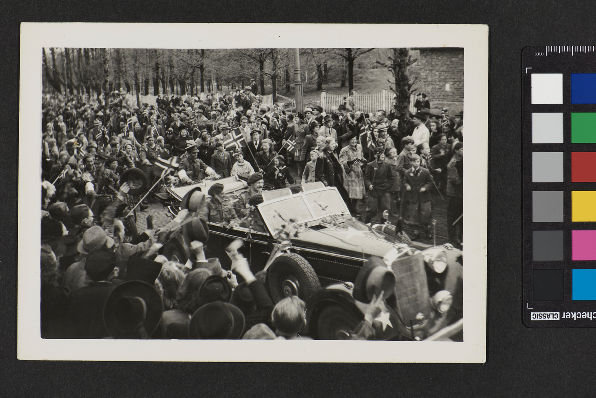 Kronprins Olav ankommer Oslo, mai 1945, i en åpen bil og er omsluttet av en folkemengde som vifter med flagg. I bakgrunnen er det trær og hvite gjerder.