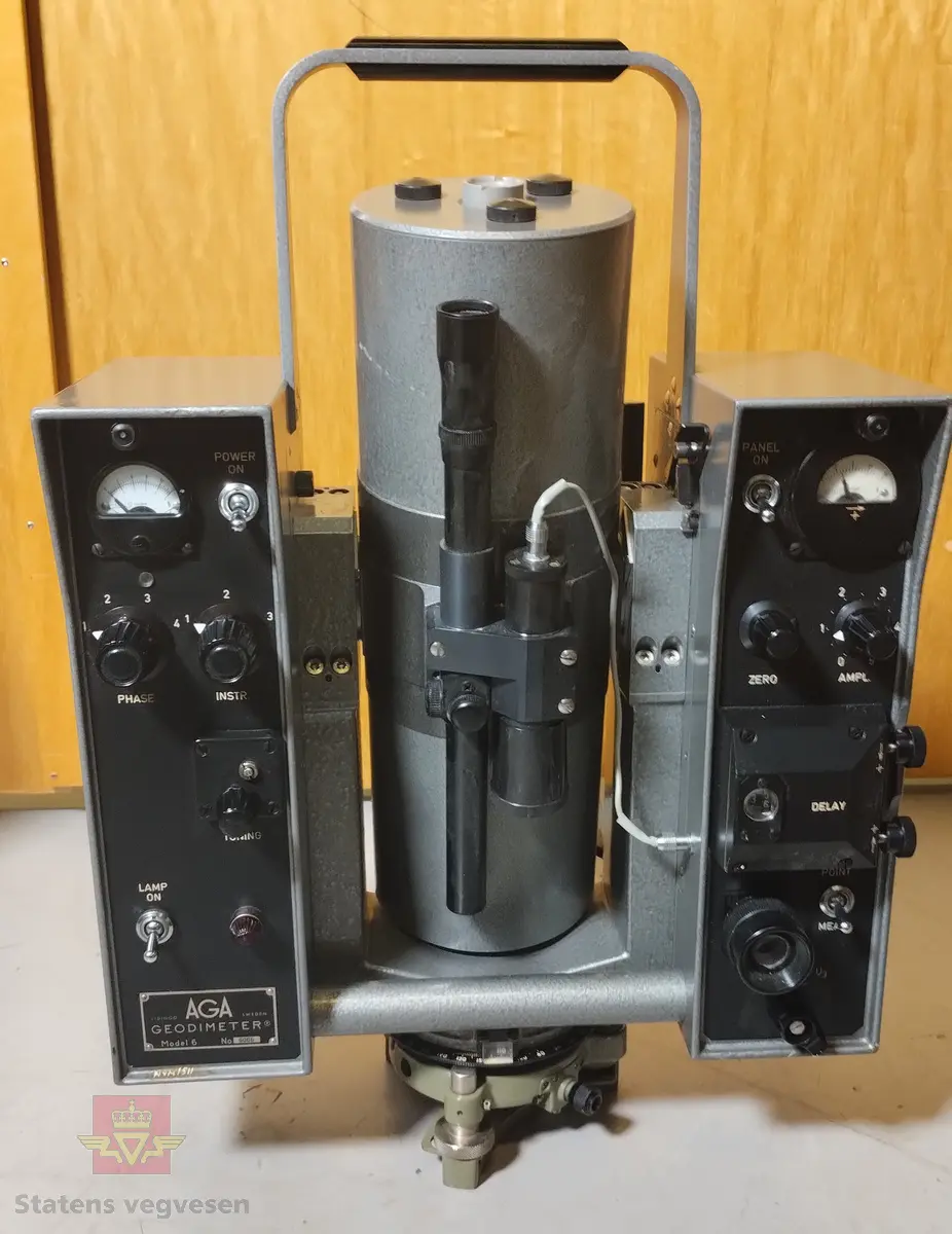 Grå og svart elektronisk avstandsmåler med tilhørende utstyr og bruksanvisning, i grå/hvit oppbevaringskasse laget av glassfiber. Merking fra produsent. Kassen er merket med lapper og klistremerker pga. frakt. På kassen er det også festet reimer slik at du kan bære kassa som en ryggsekk. 

Tilbehør som hører med er: 
- Kabel, svart og gul, med en rød og svart batteriklemme for tilkobling av batteri.
- Grå lærreim, 140 cm lang med blank spenne.
- Hengelås av messing med blank nøkkel. Lagd i England. Brukes til låsing av oppbevaringskassa.
- Håndbok med blå perm for AGA Geodimeter.
- Mappe med tabeller og papirer for AGA Geodimeter
- Grønne plastmapper med måleskjemaer for AGA Geodimeter

