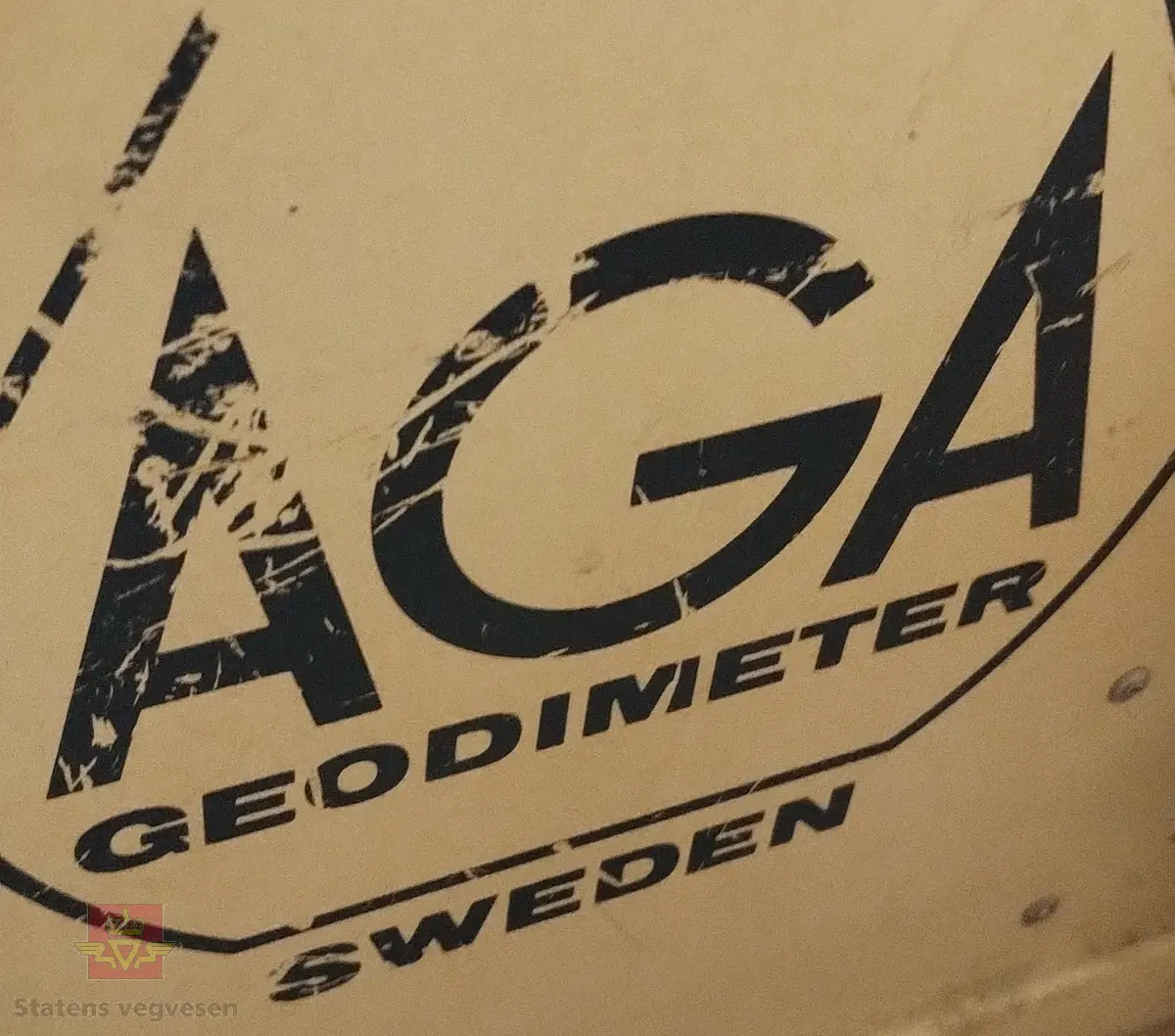 Grå og svart elektronisk avstandsmåler med tilhørende utstyr og bruksanvisning, i grå/hvit oppbevaringskasse laget av glassfiber. Merking fra produsent. Kassen er merket med lapper og klistremerker pga. frakt. På kassen er det også festet reimer slik at du kan bære kassa som en ryggsekk. 

Tilbehør som hører med er: 
- Kabel, svart og gul, med en rød og svart batteriklemme for tilkobling av batteri.
- Grå lærreim, 140 cm lang med blank spenne.
- Hengelås av messing med blank nøkkel. Lagd i England. Brukes til låsing av oppbevaringskassa.
- Håndbok med blå perm for AGA Geodimeter.
- Mappe med tabeller og papirer for AGA Geodimeter
- Grønne plastmapper med måleskjemaer for AGA Geodimeter

