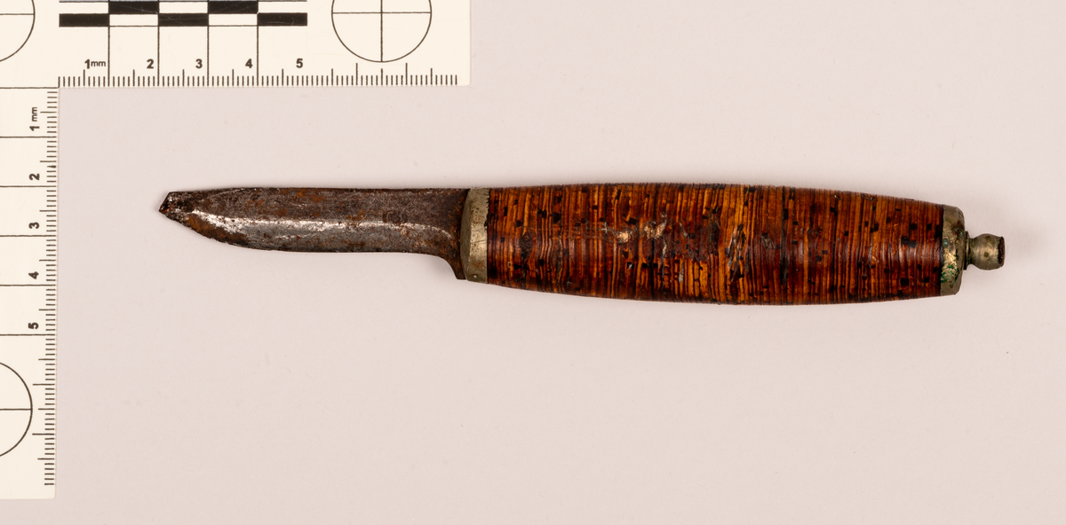 Kniv med skaft. Skaftet har hådsmidd metall i begge endene. Bakre ende av skaftet har knapp. Skaftet består av neverflak, som er slipt slik at kantene på neverflakene utgjør en flate som hånden kan omslutte. Knivbladet er slitt ned med omlag en fjerdedel av bladets opprinnelige høyde. Kniven er forseggjort og er et pent arbeid.