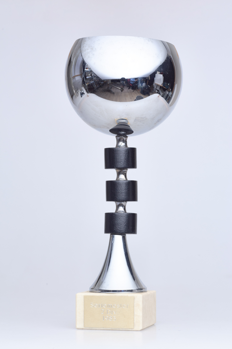 Pokal med halvrund topp, tre svarte runde ringer rundt stetten og en fot i hvit sten. Innskriften er "Seriemester 2. div. 1985"