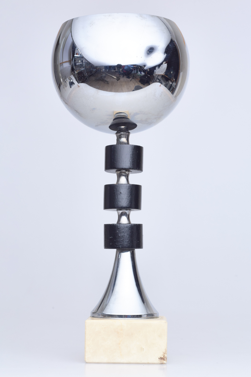 Pokal med halvrund topp, tre svarte runde ringer rundt stetten og en fot i hvit sten. Innskriften er "Seriemester 2. div. 1985"