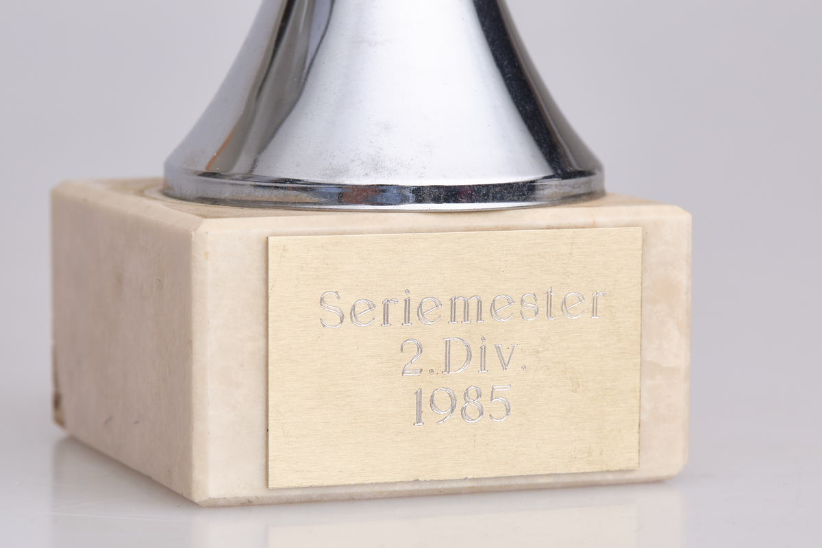 Pokal med halvrund topp, tre svarte runde ringer rundt stetten og en fot i hvit sten. Innskriften er "Seriemester 2. div. 1985"